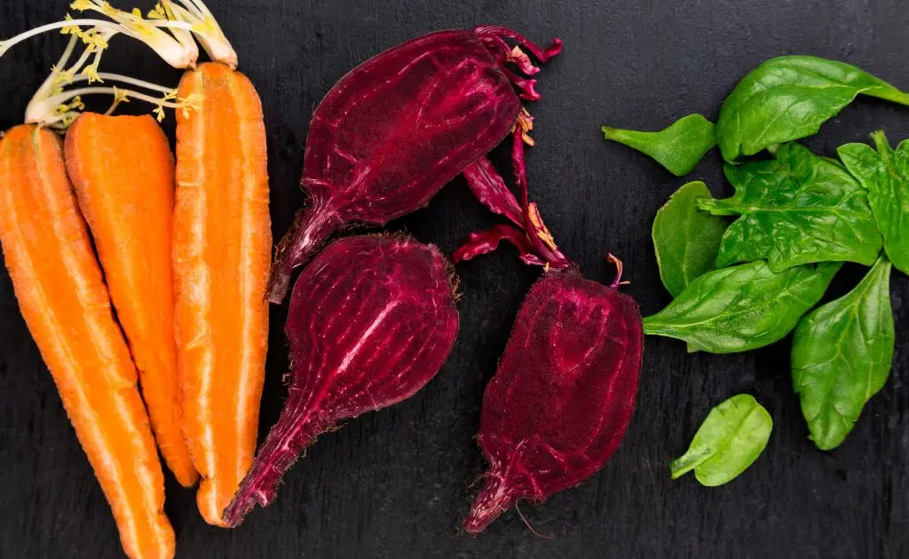 A vibrant image showcasing fresh carrots with green tops, dark red beetroots, and bright green spinach leaves arranged on a dark surface. Shows what you should eat to minimize hair problems.